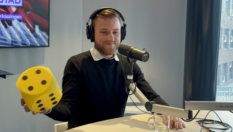 Man met koptelefoon in opnamestudio houdt een grote gele dobbelsteen vast; microfoon en tv-scherm op de achtergrond.
