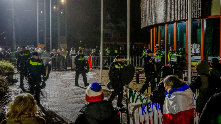 Politieagenten staan voor een gebouw; een menigte met borden bevindt zich achter dranghekken.