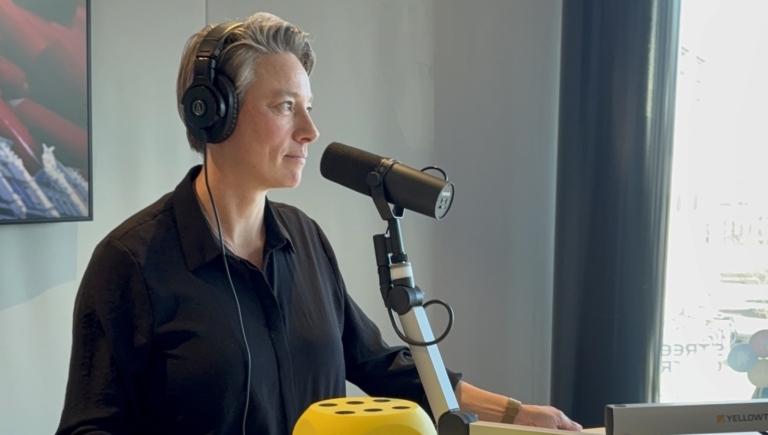 Persoon met koptelefoon spreekt in een microfoon; een grote gele dobbelsteen ligt op de tafel.