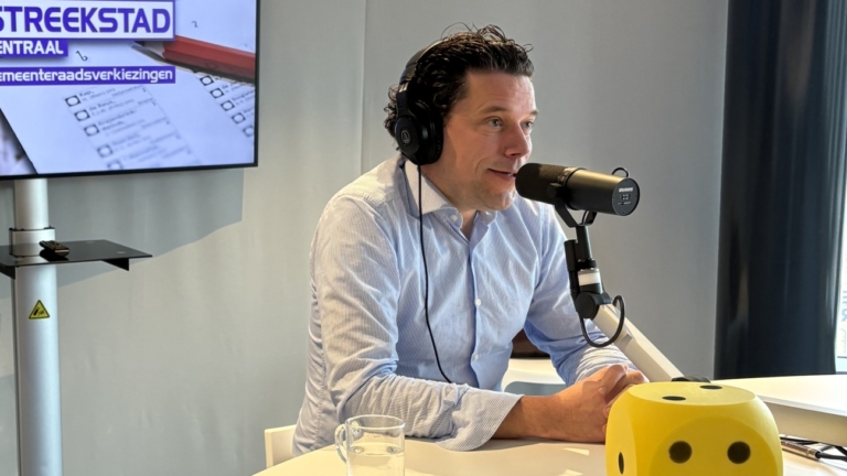 Man in radiostudio spreekt in microfoon, met scherm dat "Streekstad Centraal" met gemeenteraadsverkiezingen toont.