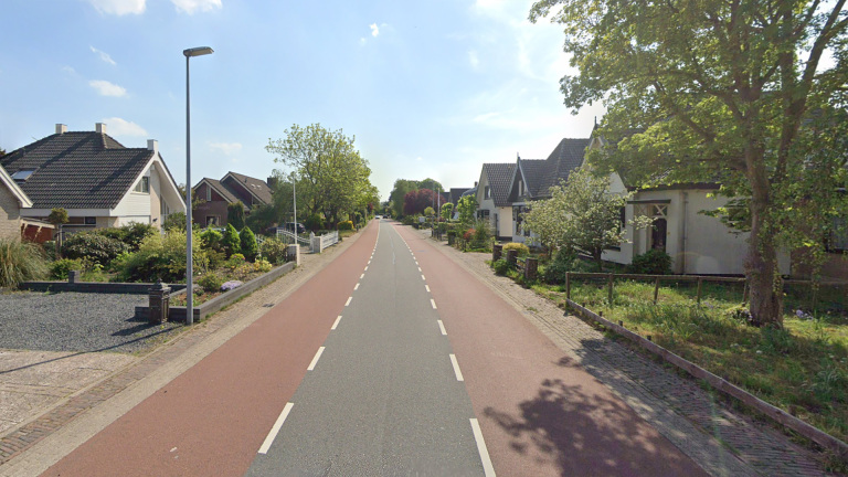 Rechte weg in een woonwijk met vrijstaande huizen, groene tuinen en bomen langs de weg onder een heldere blauwe lucht.