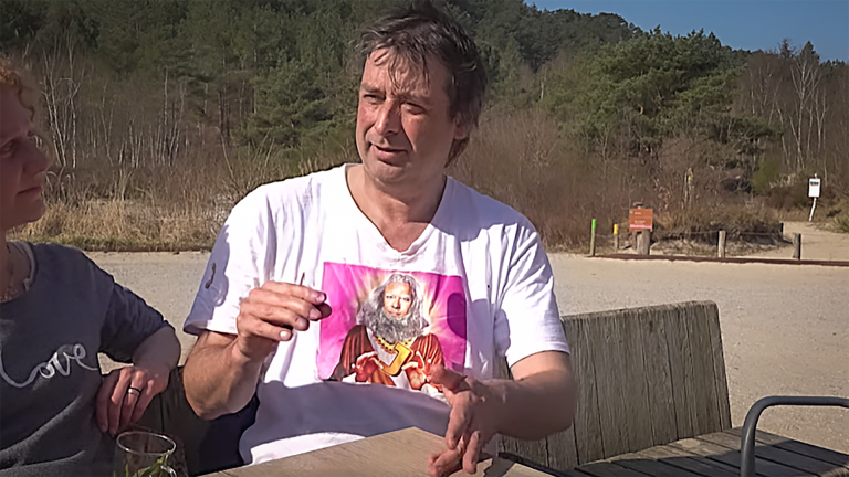 Twee personen zitten aan een tafel buiten, met een bosrijke omgeving op de achtergrond. Eén persoon draagt een opvallend T-shirt.