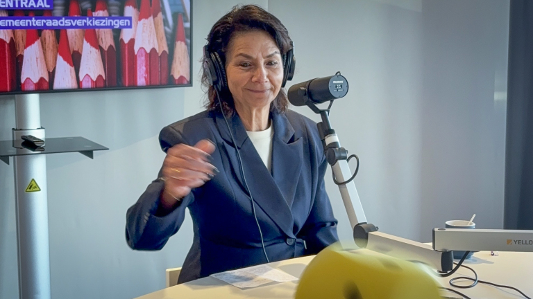 Vrouw met koptelefoon en microfoon in een studio, met op de achtergrond een scherm met verkiezingsaankondiging.