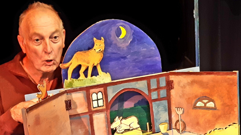 Man voert een poppenspel op met decor van een huis en een papieren wolf onder een nachtelijke hemel.