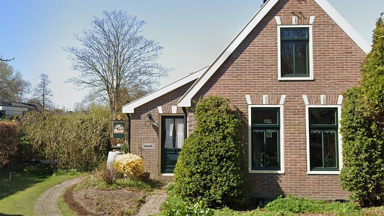 Bakstenen huis met groene deuren en ramen, omringd door een tuinpad, struiken en een bord met "Theater".