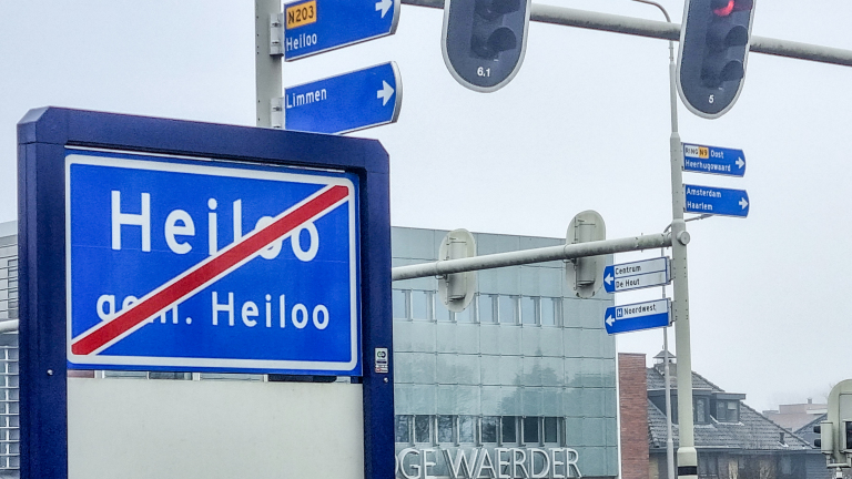 Verkeersbord met doorgestreepte naam Heiloo, met bewegwijzering voor Limmen, Heerhugowaard, Amsterdam en Haarlem op de achtergrond.