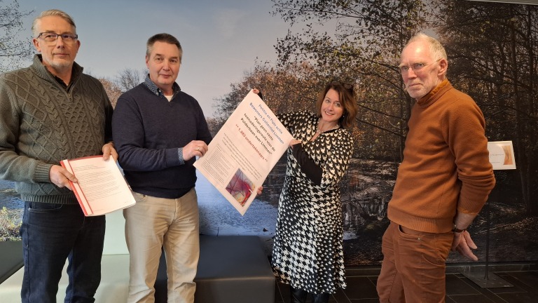 Vier mensen poseren met een groot document in een kamer met een bosachtergrond op de muur.