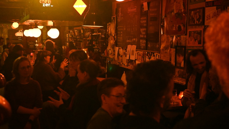 Mensen praten en drinken in een sfeervol verlichte bar met posters aan de muur.