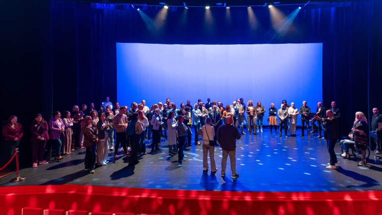 Groep mensen verzameld op een verlicht podium in een theateromgeving, omringd door blauwe achtergrondverlichting.