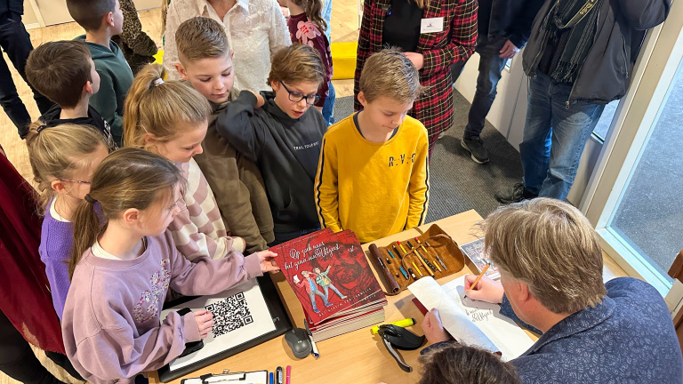 Kinderen verzamelen zich rond een auteur die een boek signeert op een tafel met stapel boeken en schrijfspullen.