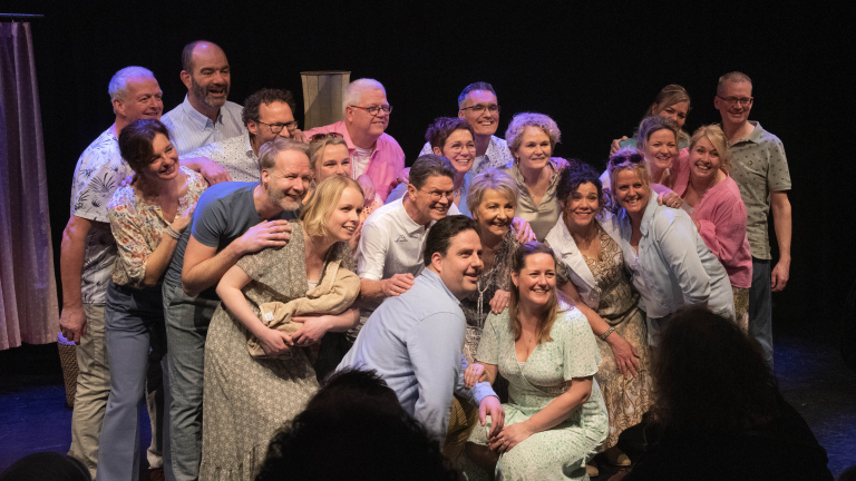 Een groep mensen poseert lachend op een podium, met theatergordijnen op de achtergrond.