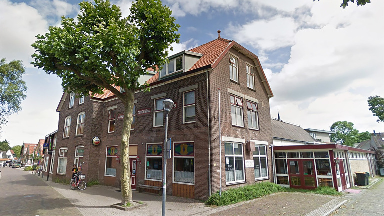 Bakstenen hoekgebouw met een rode pannendak en ramen met kleine rode zonnekleppen, omgeven door een straat met bomen en een stoep. Er staat een fiets geparkeerd voor het gebouw.