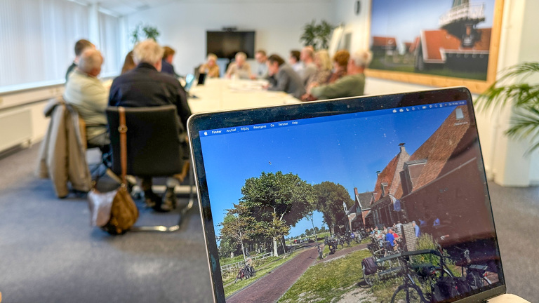 Vergaderruimte met groep mensen rond een tafel, op de voorgrond een laptop met foto van een dorp onder een blauwe lucht.