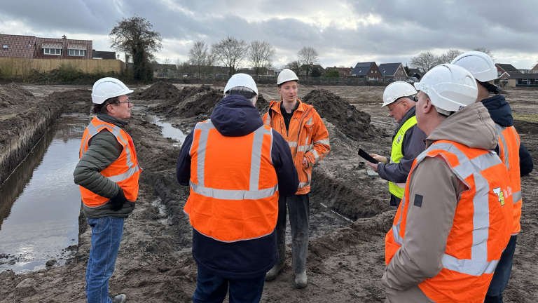 Groep bouwvakkers in reflecterende kleding en helmen overlegt op een modderige bouwplaats onder een bewolkte hemel.
