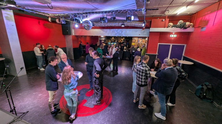 Mensen staan in groepjes te praten in een zaal met een rode muur en een bar op de achtergrond. Er hangt een discobal aan het plafond.