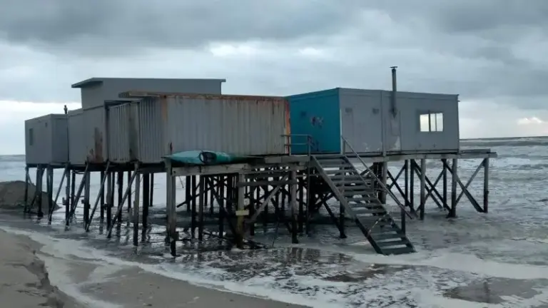 Verhoogde gebouwen op palen aan zee met bewolkte lucht op de achtergrond.