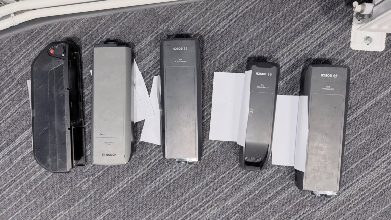 Vijf Bosch PowerPack 500 accu's liggen op een grijze gestreepte vloerbedekking, deels op witte papieren.