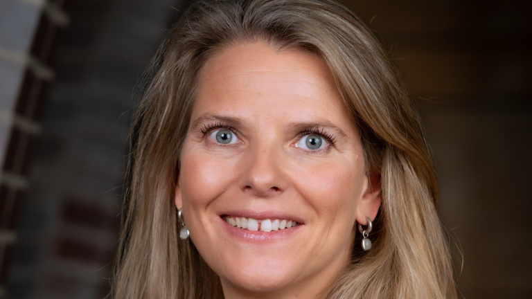 Vrouw met lang blond haar en glimlach, draagt pareloorbellen.