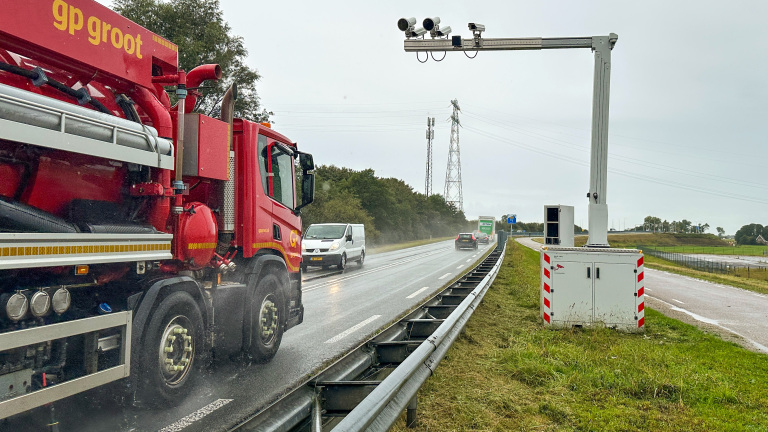 Een rode vrachtwagen rijdt op een natte snelweg langs een verkeerscamera-installatie, met meerdere voertuigen zichtbaar op de achtergrond onder een bewolkte hemel.