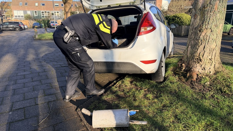 Agent onderzoekt de kofferbak van een witte auto naast een boom, met een auto-onderdeel op de grond.