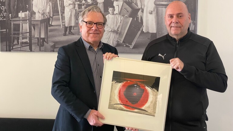 Twee mannen houden een kunstwerk met een rood en zwart abstract ontwerp vast, voor een zwart-wit fotoachtergrond.