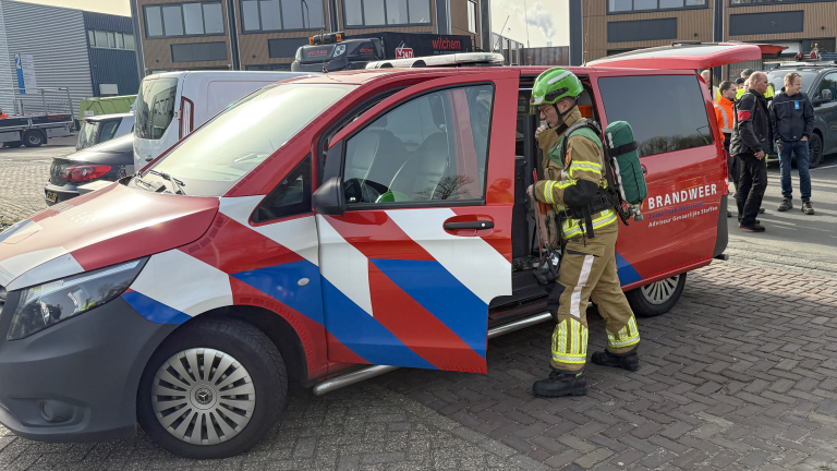Brandweerman in uitrusting staat bij een brandweerwagen met geopende deur, omgeven door andere mensen en voertuigen.