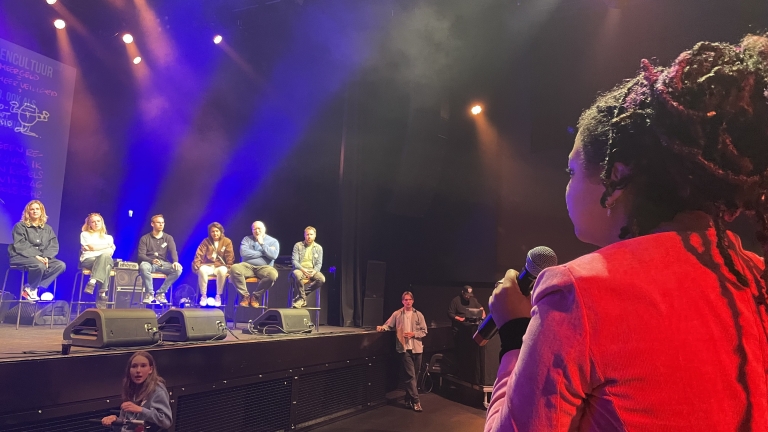 Vrouw met microfoon spreekt tot panel met zes personen op een podium in een zaal met gekleurde verlichting.