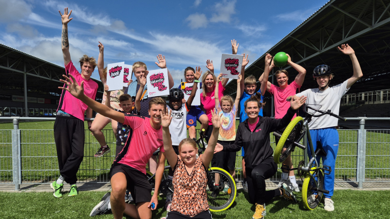 Een groep mensen in sportkleding die enthousiast zwaait en poseert in een stadion, met borden en een fiets.