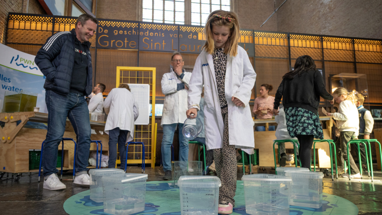 Een meisje in een witte jas giet water in een bak tijdens een wetenschapsactiviteit, omringd door volwassenen en kinderen.
