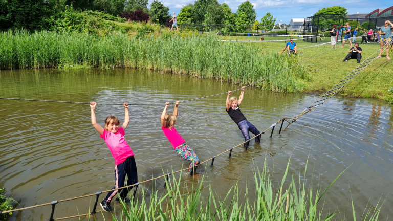 Drie kinderen hangen aan een touwbrug boven een vijver, terwijl omstanders toekijken op de achtergrond.