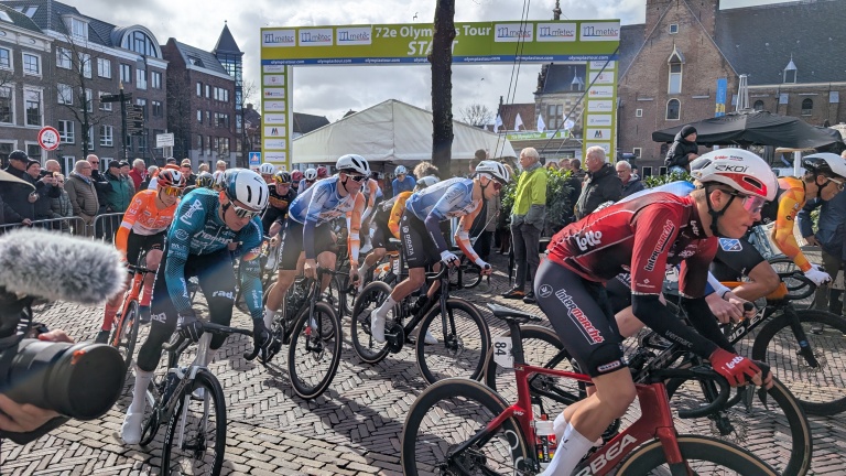Wielrenners starten bij de 72e Olympia's Tour onder een boog, met toeschouwers op de achtergrond.