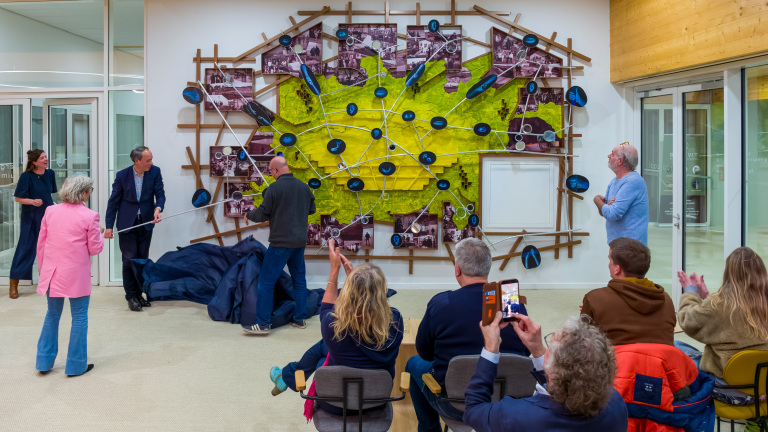 Mensen onthullen en bewonderen een groot kunstwerk aan de muur met foto's, gele kleurvlakken en verbindingslijnen.