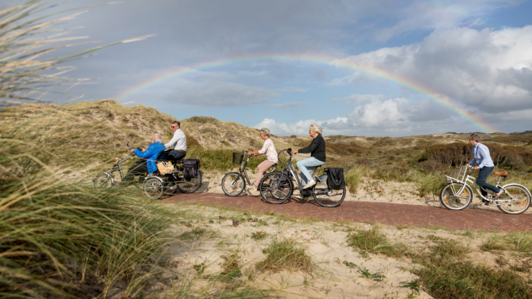 Mensen fietsen langs een pad in de duinen met een regenboog aan de lucht.