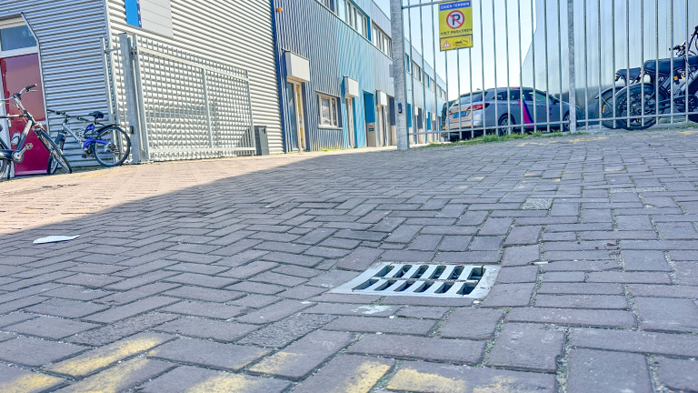 Een industrieterrein met fietsen, een afvoerrooster in de bestrating en een bord met "Niet Parkeren" achter een hek.