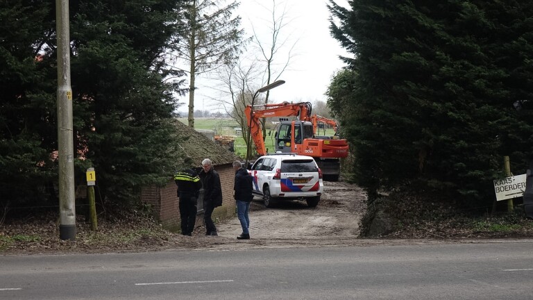 Politieagenten en een politiewagen staan bij een bouwplaats met een graafmachine, omgeven door bomen.