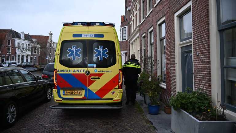 Een ambulance en een politieagent bij een bakstenen gebouw in een smalle straat, met geparkeerde auto's en grachtenhuizen op de achtergrond.