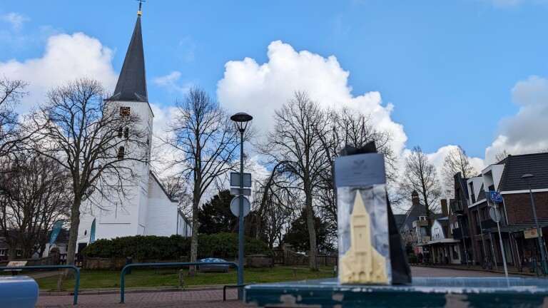 Een witte kerk met een hoge, puntige toren omringd door kale bomen en een helderblauwe lucht, met een verpakking vooraan met een miniatuurversie van de kerk.