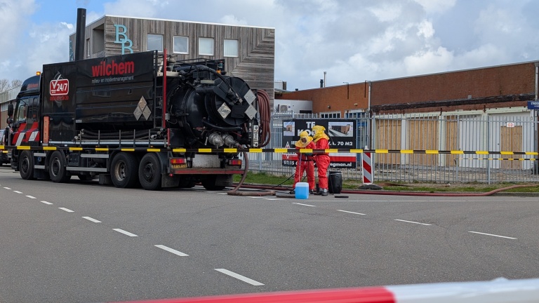 Vrachtwagen van Wilchem en twee personen in beschermende kleding op straat bezig met een klus, afgezet met lint.