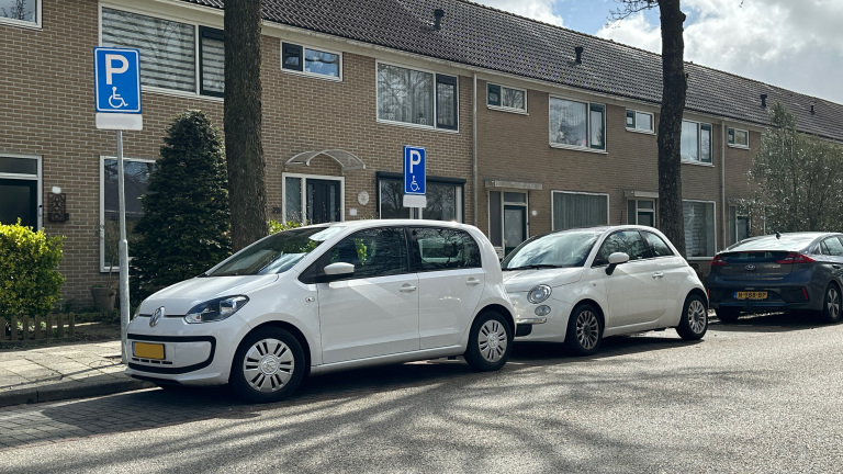 Witte auto's geparkeerd in een rij op straat, naast een bord voor gehandicaptenparkeerplaatsen, voor een rijtjeshuis.