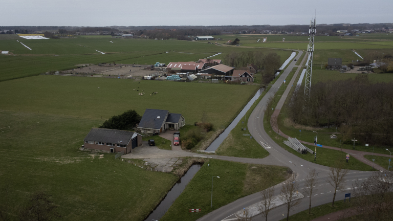 Landelijk gebied met boerderij, weilanden, een weg en een zendmast, omgeven door kanalen en verspreide bomen.