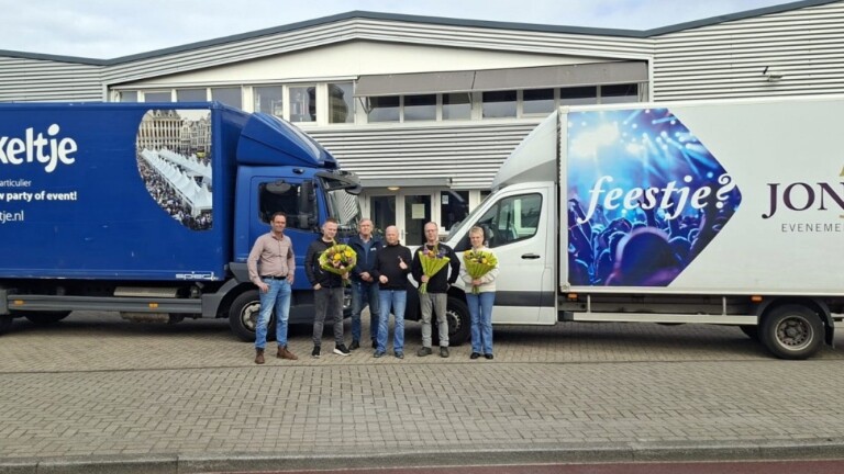 Zes mensen staan voor twee vrachtwagens, drie van hen houden boeketten vast; de voertuigen hebben reclameteksten.