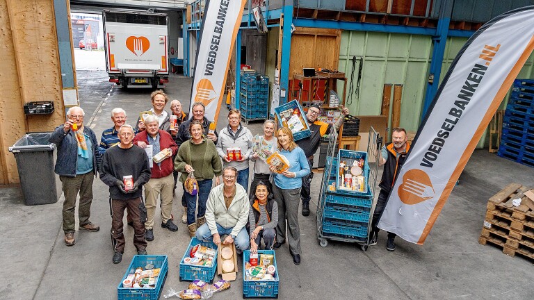 Groep vrijwilligers poseert met voedselpakketten in een magazijn met Voedselbank-vlaggen op de achtergrond.
