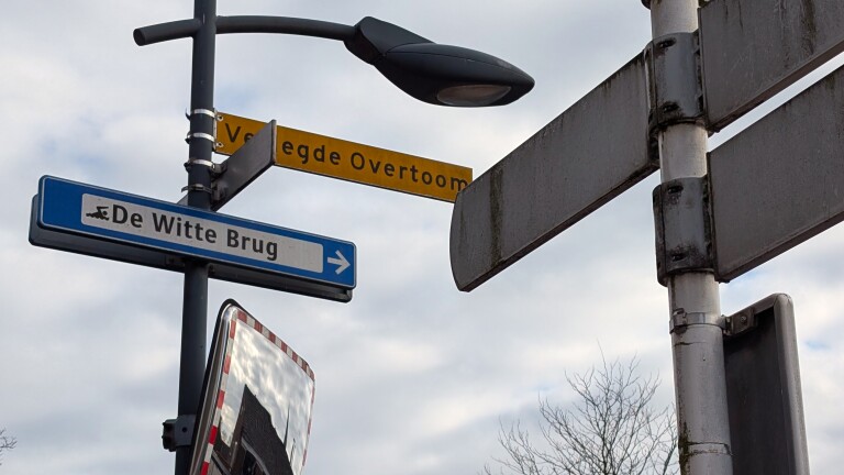 Straatnaamborden met "Voetwegde Overtoom" en "De Witte Brug" bij een lantaarnpaal onder een grijze lucht.
