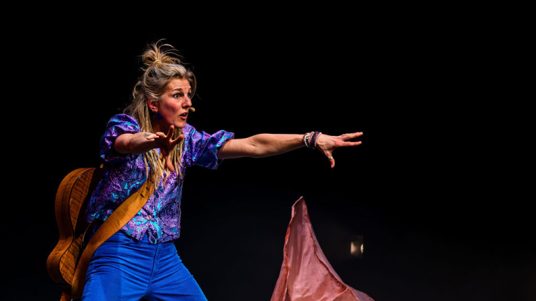 Vrouw in kleurrijke outfit met gitaar op de rug, gebaart expressief op een podium tegen een donkere achtergrond.