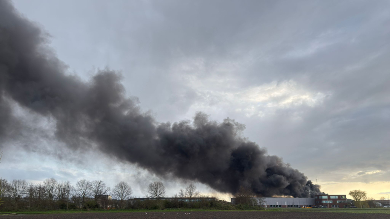 Een grote zwarte rookpluim stijgt op uit een gebouw in een industriegebied onder een bewolkte hemel.