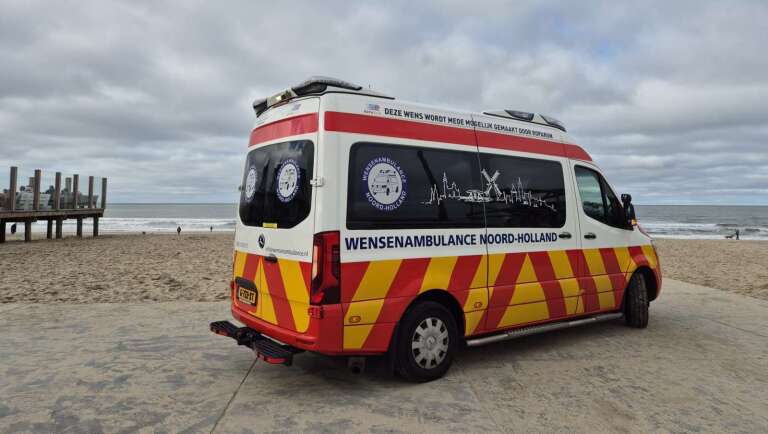 Een Wensambulance Noord-Holland geparkeerd bij een strand met deels bewolkte hemel op de achtergrond.