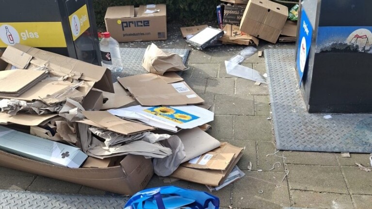 Afval bestaande uit karton en papier ligt verspreid bij een glascontainer op een stoep, samen met een lege fles en doosjes.