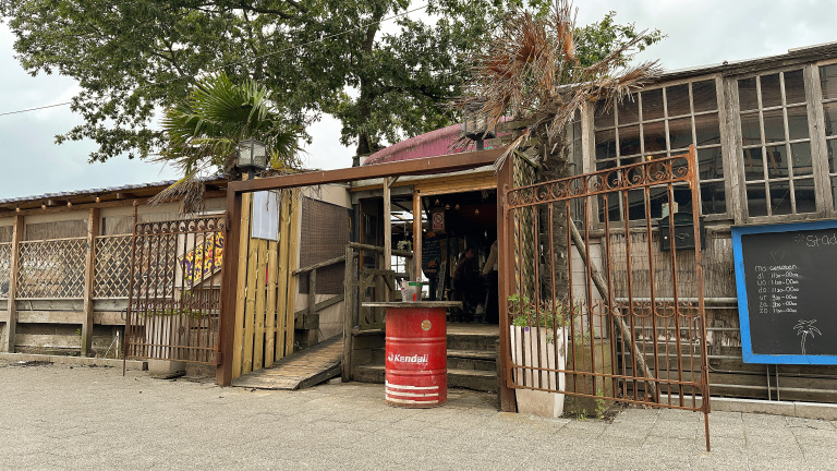 Entree van een houten gebouw met een openstaande ijzeren poort en een rood olievat als tafel. Er hangt een uithangbord met openingstijden naast de ingang. Boven de poort is een palmboom zichtbaar.