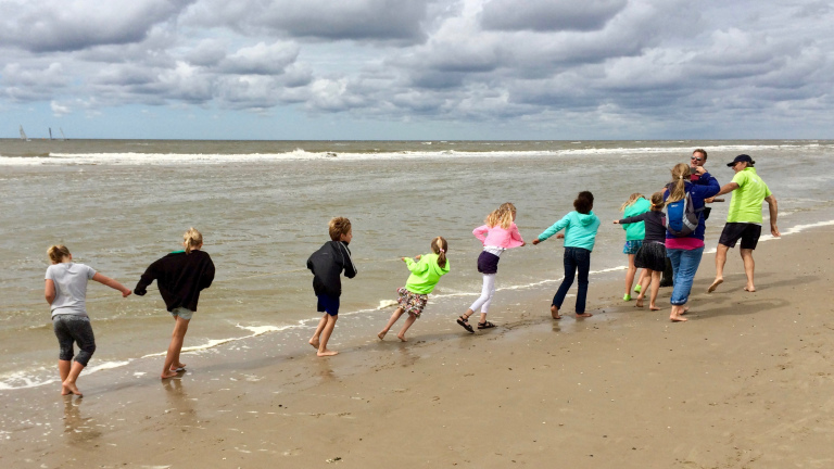 Kinderen en volwassenen spelen hand in hand op het strand langs de kustlijn onder een bewolkte hemel.