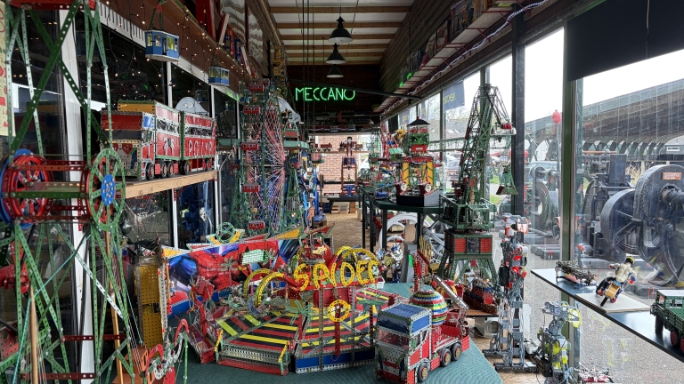 Binnenruimte vol met kleurrijke Meccano-modellen, waaronder miniatuurattracties en voertuigen, met op de achtergrond een helder verlicht "Meccano" bord.
